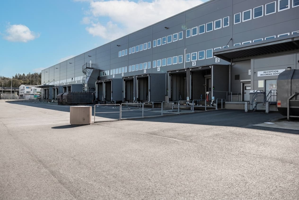 A large warehouse loading bay with several dock doors, a lorry trailer, metal barriers, and a reception sign under clear skies.