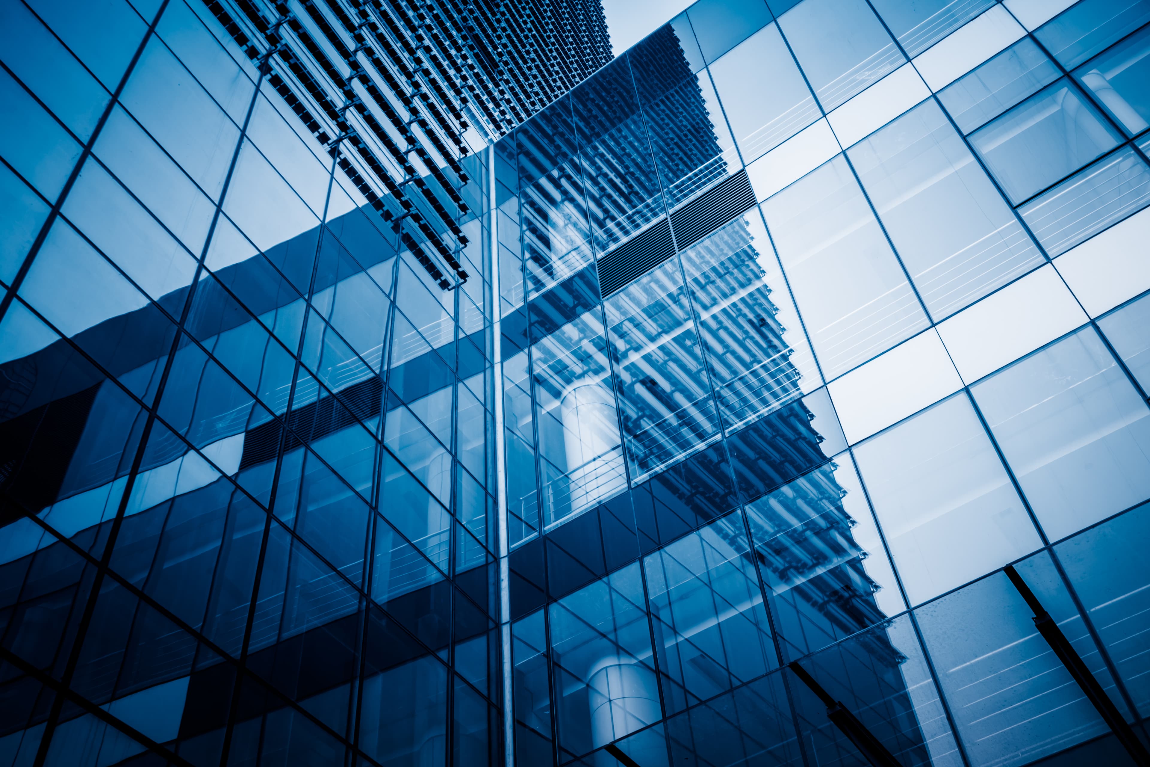 Modern glass building façade reflecting surrounding architecture, forming a pattern of blue-tinted windows and geometric lines.